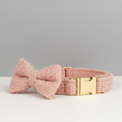 A pink dog bow resting against a collar of the same material and style on a white table with a gray wall behind it, facing backwards